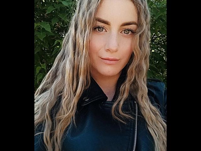 BlondyOneSex, con i capelli biondi mossi, gli occhi verdi e un sorriso discreto, indossa una giacca di pelle nera, guarda direttamente la fotocamera e ha un viso carino e sensuale..