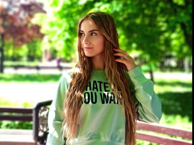 BlondyOneSex con cabello castaño largo y ondulado, sudadera verde, mano en el cabello, de pie junto a un banco rosa, fondo de parque verde, mirando hacia un lado con una ligera sonrisa..