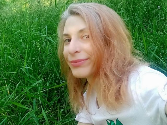 Une femme séduisante avec des cheveux bruns clairs ondulés, des lèvres roses et un léger sourire, portant une chemise blanche, debout dans de hautes herbes vertes. Ses seins sont petits, et elle est mignonne..