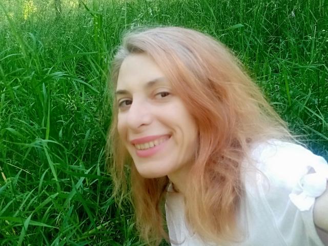 ASexyLady with pinkish-blonde hair, smiling, white shirt, grassy background, cute, soft face, small nose, pink lips, light skin, natural light, casual selfie.