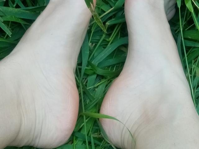 ASexyLady's pale feet with pink soles are lying on green grass, showing soft, slightly wrinkled skin with a few blades of grass in between.