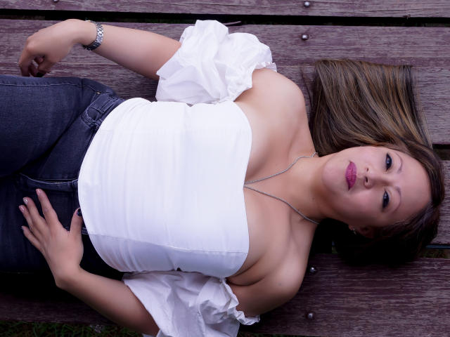 ChristinMoore is lying on wooden planks, wearing a white off-shoulder top, blue jeans, and pink lipstick. Her tits are visible, and she has a silver necklace.