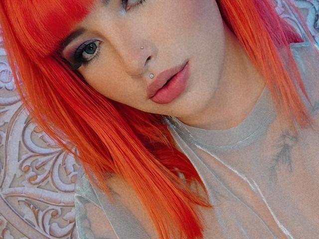 BrunnaCatie, com cabelo laranja vibrante, olhos azuis, piercings no nariz e pele clara, vestindo uma blusa cinza transparente, exibindo uma expressão suave e levemente com a boca entreaberta. Seus seios estão parcialmente visíveis..