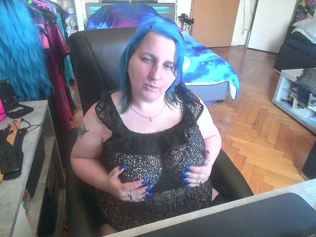 DachenHot met blauw haar, een zwarte kanten jurk, die haar grote borsten vasthoudt, zittend in een zwarte stoel, met een houten vloer, en een kleurrijk bed op de achtergrond, met een lichte glimlach..