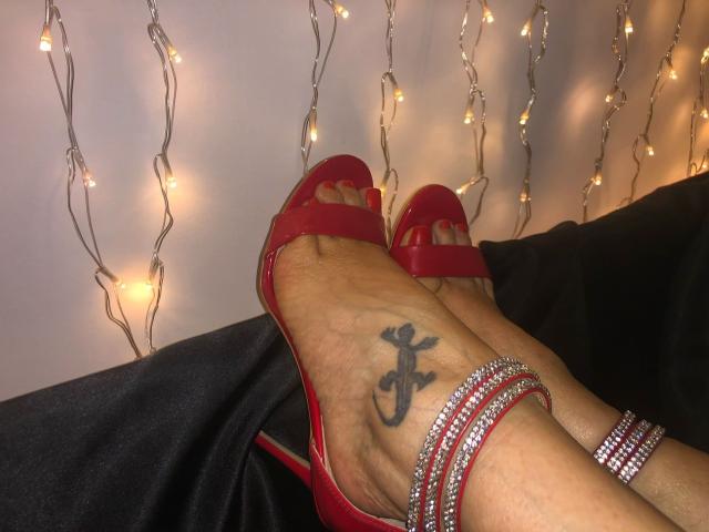 SiPassioneX's red high-heel sandals with red toenails, ankle bracelet, and black anchor tattoo on her foot, resting on black fabric. Warm fairy lights in the background.