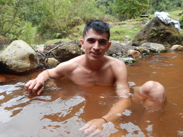 Um homem latino, chamado Andres, sem camisa, com cabelo preto curto, pequenas tatuagens e pele morena, está em um rio lamacento, com o joelho levantado, sorrindo, o braço direito na água e o braço esquerdo em uma rocha..