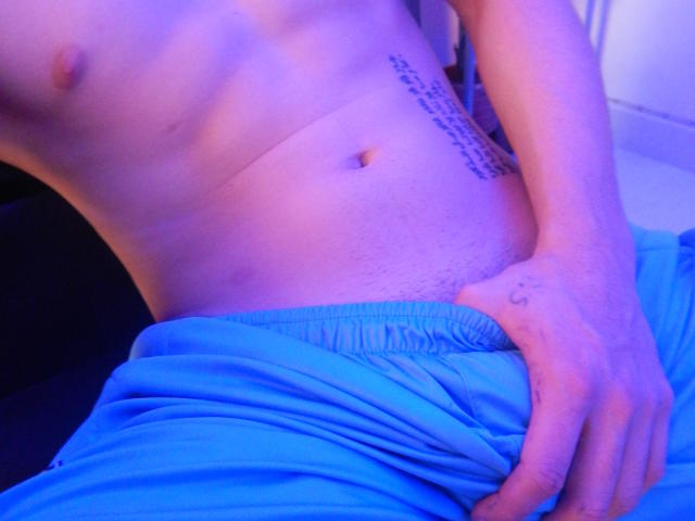 O torso nu de LatinAndres, iluminado em tons de roxo, com uma tatuagem no lado direito, calças azuis puxadas para baixo, revelando uma mão cobrindo seu pênis peludo e não circuncidado..