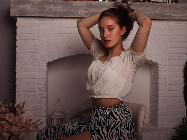EvaPink with arms up, wearing a white puff-sleeve crop top and zebra-striped pants, standing in front of a white brick fireplace, looking slightly to the side.