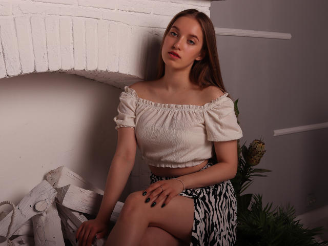 EvaPink with pale skin, brown hair, small boobs, wearing a white off-shoulder top and zebra-striped skirt, sitting on a white wooden structure, looking slightly to the side.