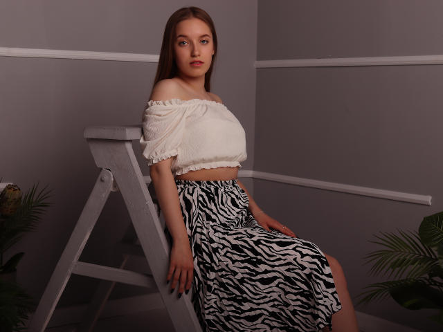 EvaPink, avec de petites seins, portant un crop top blanc à une épaule et une jupe à motifs zébra, assise sur une échelle blanche dans une pièce avec des plantes vertes..