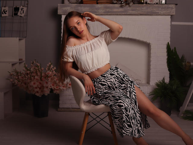 EvaPink with long brown hair, off-shoulder white top, and zebra-striped skirt, sitting on a chair, hand in hair, legs crossed, showing her tits and a sexy smile.