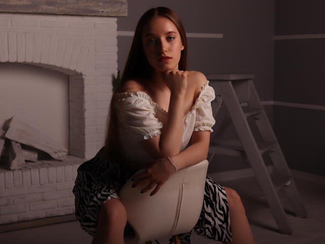 EvaPink, avec de longs cheveux bruns, un haut blanc à épaules dénudées et une jupe à motifs zébra, assise sur une chaise blanche, tenant ses seins d'une main, avec une expression sérieuse..