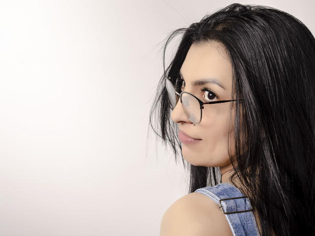 JanetteRider with long black hair, wearing glasses, and a denim sleeveless top, looking over her shoulder with a slight smile, showing her brown eyes and subtle makeup.
