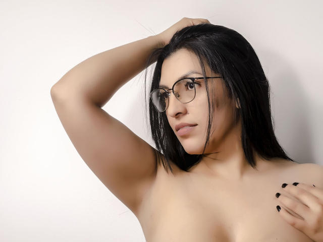 JanetteRider with black hair, glasses, and black nails, topless, arm raised, looking side, light skin, small tits, subtle smile, white background.