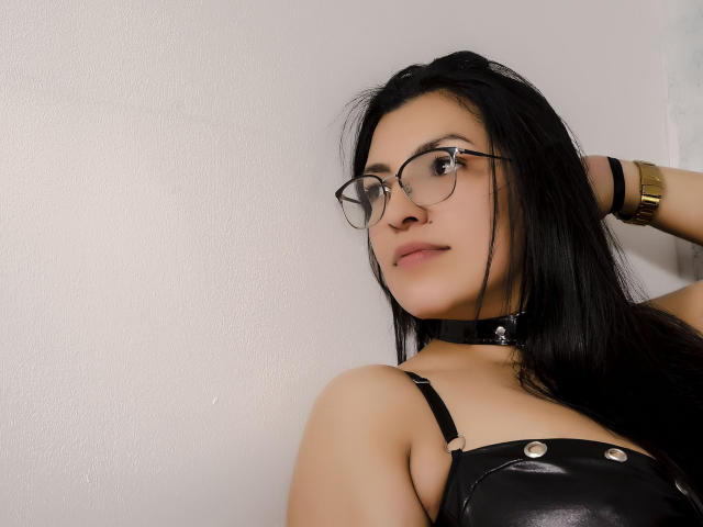 JanetteRider, con cabello negro, gafas, collar negro y top de cuero negro, mostrando sus senos, con el brazo levantado, pulsera dorada, fondo de pared blanca, con una ligera sonrisa..
