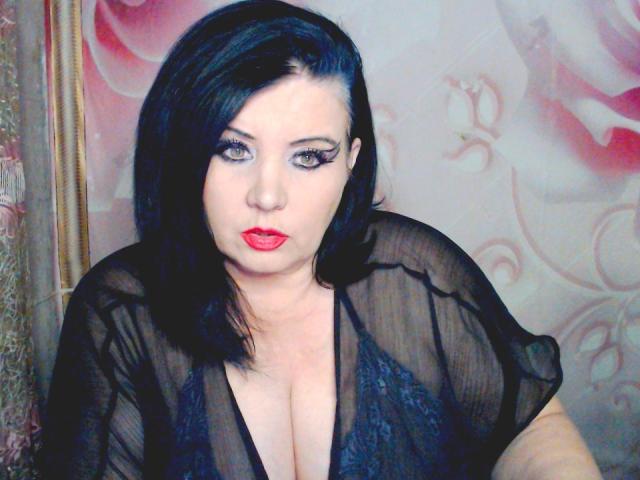 KamilaDream, com cabelo preto, batom vermelho e uma blusa preta transparente que mostra seios grandes, com uma expressão séria, usa delineador marcante e está centralizada no quadro..