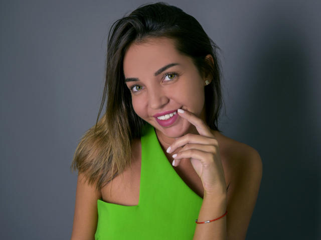 LettieSweet with medium tits, wearing a bright green halter top, has brown hair, green eyes, and a cute smile, touching her finger to her lips, with a red bracelet.