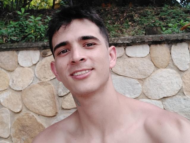 LatinAndres tem cabelo curto e escuro, olhos castanhos e uma pequena tatuagem no pescoço. Ele está sem camisa, sorrindo com um pequeno espaço entre os dentes, e tem uma pele clara..