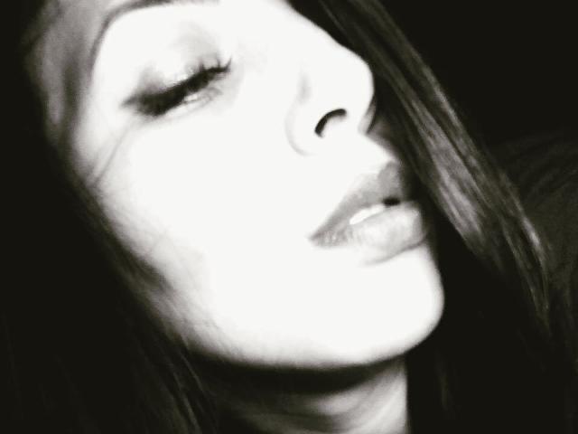 El rostro de EveNetty en un selfie en blanco y negro, con los ojos entreabiertos, el cabello largo y oscuro cubriendo un ojo, labios ligeramente entreabiertos y brillantes, una luz suave en su mejilla, una sonrisa suave..
