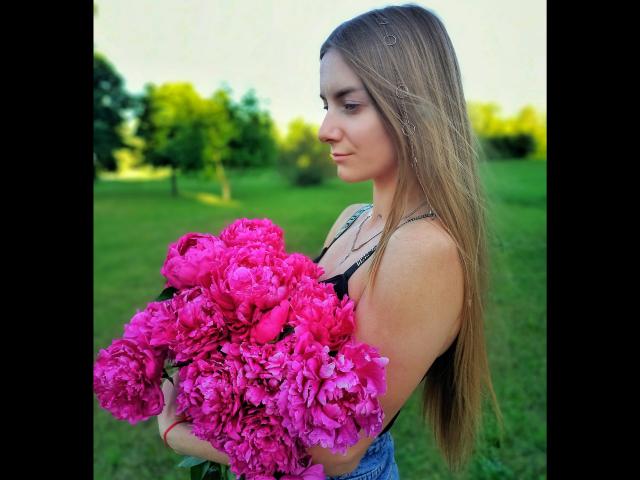 BlondyOneSex, com cabelos longos e lisos, segurando um grande buquê de peônias rosa vibrante, vestindo uma blusa preta, em pé em um campo verde, sorrindo suavemente..