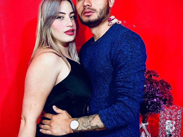 JulianYDianne, com cabelo loiro, vestido preto e batom vermelho, está em pé ao lado de um JulianYDianne de barba, vestindo um suéter azul, segurando-a contra uma parede vermelha..