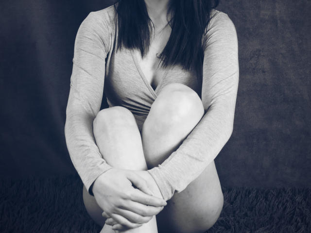 PassionGirlX, met haar knieën opgetrokken, draagt een strak, lang袖 shirt dat haar kleine borsten en een subtiele decolleté laat zien, en zit op een donker, pluizig tapijt..