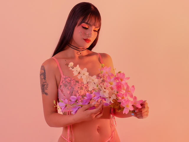 Gaby Durand, con biancheria intima rosa, tiene in mano fiori rosa e viola, ha tatuaggi sul braccio destro e indossa una collana nera. Ha capelli lunghi e scuri e rossetto rosso..