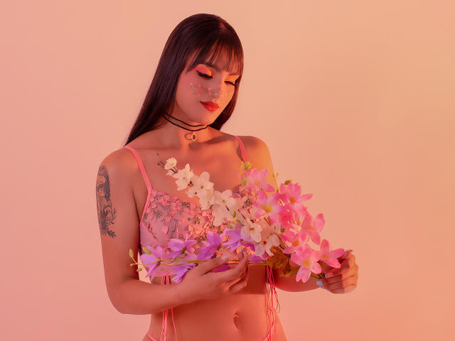 Gaby Durand mit langen, schwarzen Haaren, rosa Dessous, die rosa und weiße Orchideen hält, Tätowierung am linken Arm, rosa Gesichtsbemalung, blickt nach unten, sanfte, rosa Beleuchtung, schwarzer Choker..