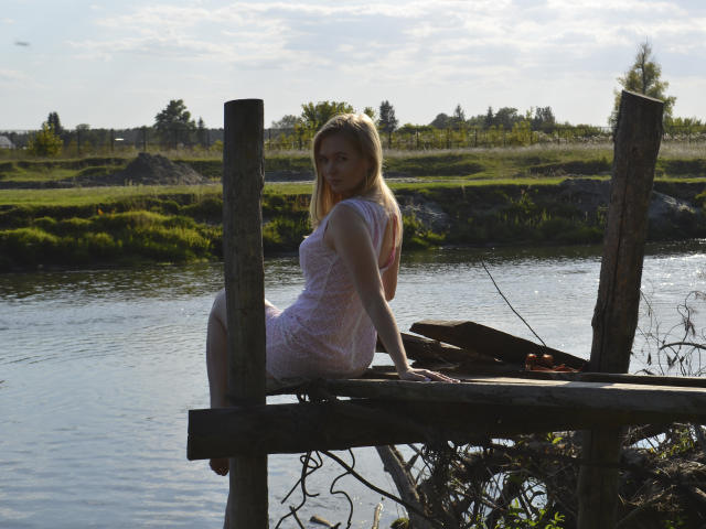 SweetAleksaX, blonde, sitting on wooden planks by a river, wearing a sheer white dress, showing her tits, looking back, sunlight, cloudy sky, rustic, calm, natural setting.