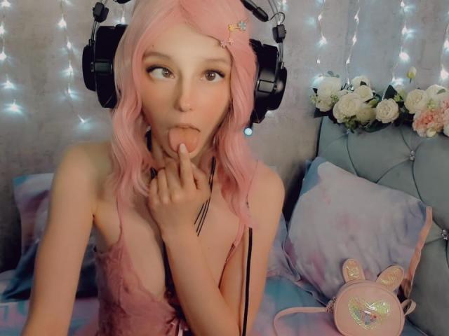 MissAstravert with pink hair, wearing headphones, sucking her finger, looking up, in a pink dress, on a bed with flowers and a bag with a cute face.