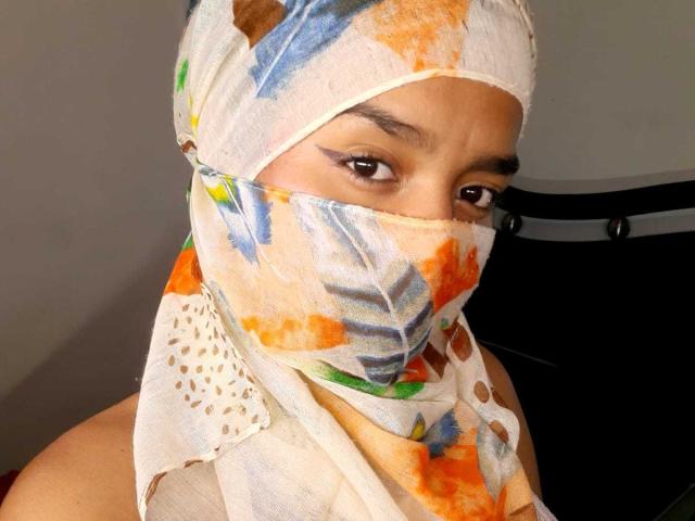 MalaLax indossa una sciarpa colorata con un motivo e una maschera che le copre il naso e la bocca, mostrando i suoi occhi marroni e le sopracciglia scure. Indossa una canotta bianca senza maniche..