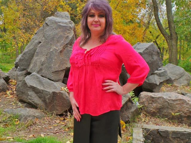 FlowerElena with medium tits, pink blouse, black pants, standing in front of large rocks, hands on hips, smiling, purple hair, autumn forest background, soft natural light.