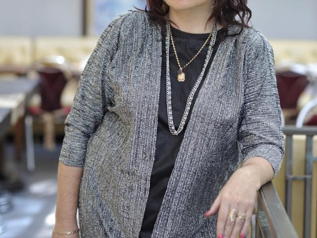 FlowerElena with gray textured cardigan, black shirt, gold necklace, and ring, slightly pregnant, standing, right hand on railing, brunette, wavy hair, blurred background.