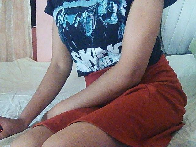 Touchie sitzt in roten Shorts und einem schwarzen T-Shirt mit einem Aufdruck auf einem weißen Bett, die Arme ruhen. Das Foto zeigt sie ab der Taille, ihre Haut ist gebräunt..