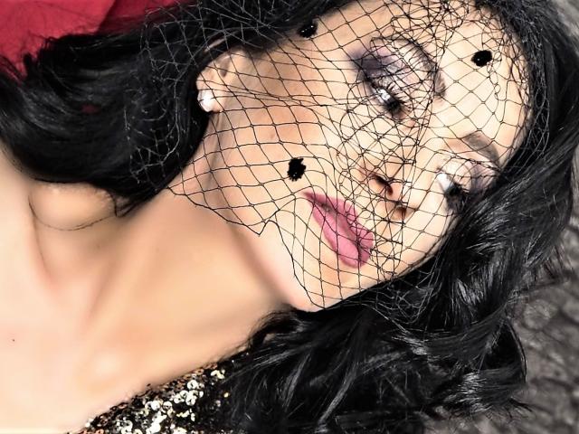 MistressTaylor's face with black wavy hair, wearing a fishnet mask covering her eyes, pink lipstick, and white earrings, against a red cloth. Her tits are partially visible.