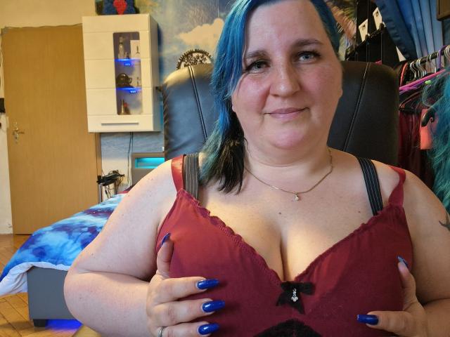 DachenHot met blauw geverfd haar, lachend, draagt een rode bh en houdt haar grote borsten vast met blauwe nagels, in een slaapkamer. Haar borsten zijn zichtbaar, ze is schattig en zelfverzekerd..