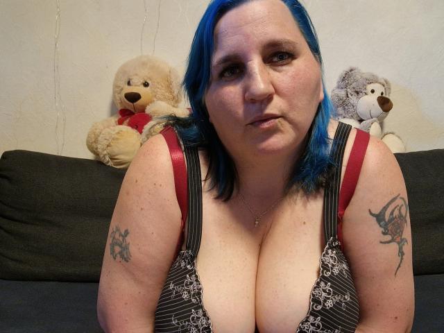 DachenHot met blauw haar, grote borsten, rode beugels van een bh, tatoeages op beide armen, zittend op een donkere bank en recht in de camera kijkend. Achter haar staan twee knuffeldiertjes..