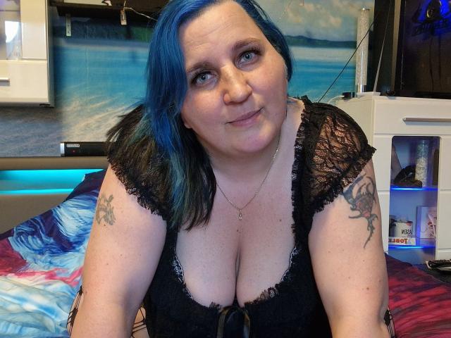 DachenHot met blauw haar, een zwarte kanten top, waarbij haar borsten zichtbaar zijn, tatoeages op haar armen, lachend, in een slaapkamer, voorovergebogen, een camera vasthoudend..