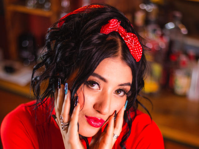 TifanyDiaz with curly black hair, red polka dot bow, red lipstick, and long blue nails, hands on face, wearing red top, seductive smile, shiny pink nails, ring.