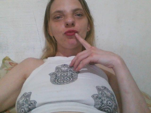 BlondyMarianne, com pele clara, cabelo loiro e batom vermelho, chupando o dedo, vestindo uma camiseta branca com estampas de hamsa em cinza, deitada..