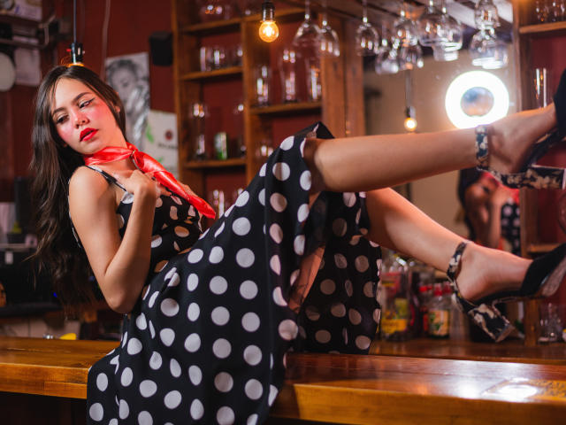 Jazmin Wells, mit einem Punktekleid, einem roten Schal und hohen Schuhen, hat ihre Beine auf einer Holzstange angelegt und zeigt ihr Gesäß in einer sexy, entspannten Pose..