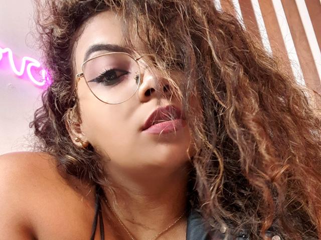 EbonyRose69 ha i capelli castano ricci, occhiali con montatura dorata e eyeliner rosa, con le labbra leggermente dischiuse, che mostrano il suo rossetto rosa. Indossa una maglietta nera e una collana dorata..