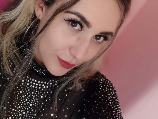 MabyLegrand has fair skin, long blonde-to-brown hair, and is wearing a black, studded top with red lipstick, looking at the camera with a slight smile.