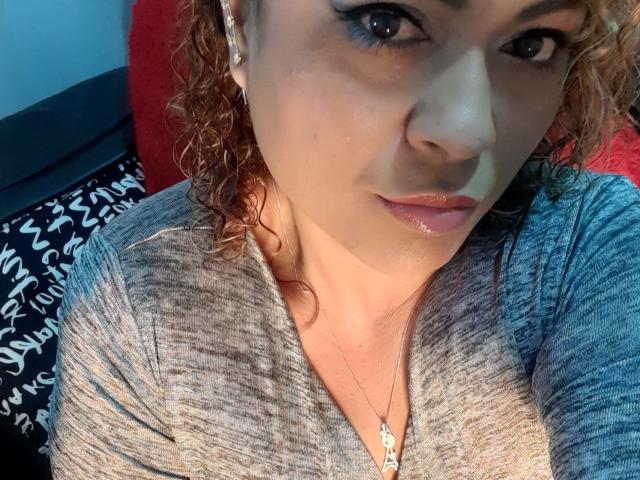 SolmaryHot with curly red hair, blue eyes, and a grey shirt, showing a slight pout with shiny lips, wearing a necklace. She's looking directly at the camera.