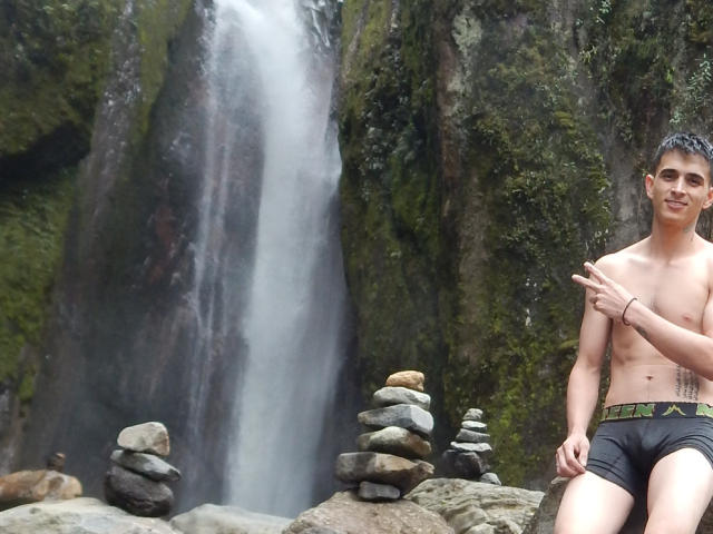 LatinAndres, sem camisa, roupa íntima preta, tatuagem no lado direito, apontando com a mão direita, em pé perto de pedras empilhadas, cachoeira ao fundo, sorrindo, cabelo curto e escuro, pele clara..