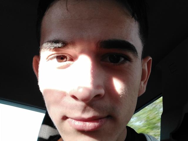 O rosto de LatinAndres, com um leve sorriso, sobrancelhas escuras e olhos castanhos, com a luz do sol iluminando parcialmente sua bochecha esquerda. Ele está vestindo uma camisa escura e sentado em um carro..