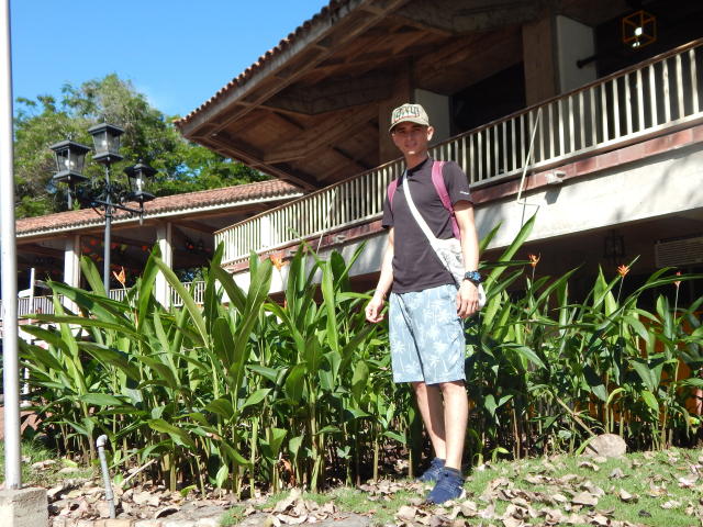 LatinAndres está em frente a plantas verdes, usando um boné, uma camisa preta, shorts azuis e tênis. Ele tem uma mochila e um relógio de pulso, em um ambiente externo ensolarado..