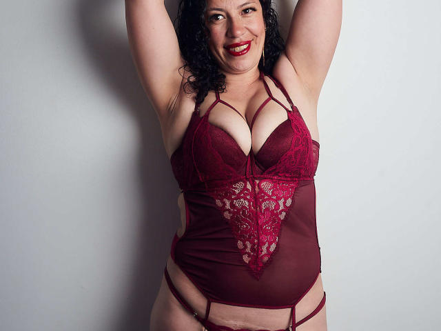 Zaly, com cabelos pretos encaracolados, batom vermelho, sorrindo, braços levantados, vestindo lingerie de renda vermelha que realça seus seios grandes e seu corpo curvilíneo, em pé em frente a uma parede branca..