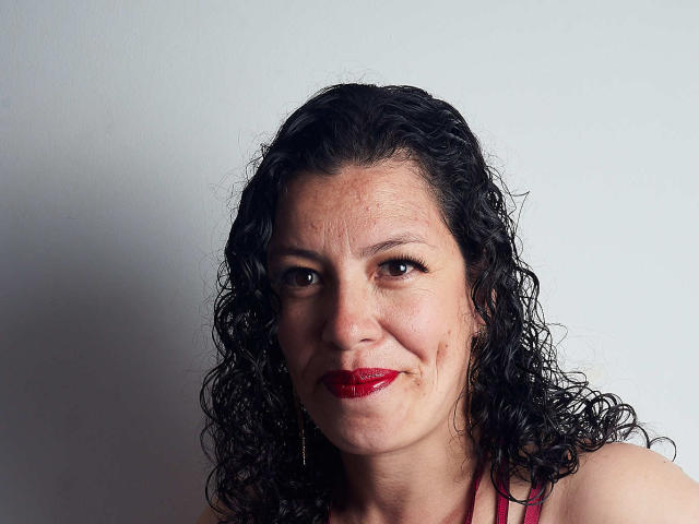 ZalyMature tem cabelo preto encaracolado, batom vermelho e um leve sorriso, com uma pequena hematoma na bochecha direita. Ela está vestindo uma blusa rosa..
