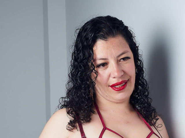 Zaly, con i capelli neri ricci, rossetto rosso e top a spalline color borgogna, sorride alla fotocamera, mostrando un seno morbido e una parte dei glutei nella parte superiore dell'immagine..